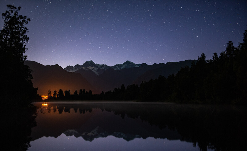 Night landscape with silhouetted mountains, forest, and a lakeside cabin reflected in still water beneath a starry sky.