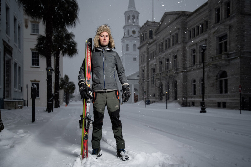 Skier standing in snowy town square holding red skis and poles, wearing winter gear.