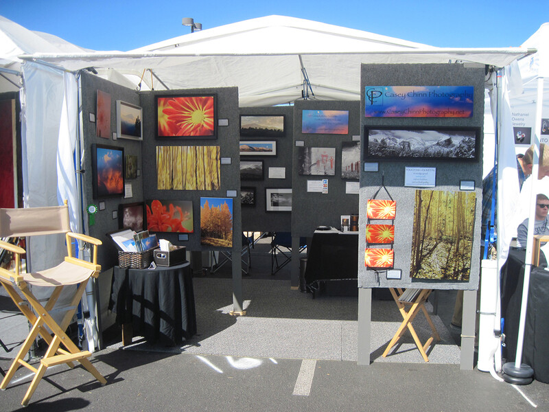 Artist's outdoor booth displaying landscape and nature photographs on black panel walls beneath white tent canopy.