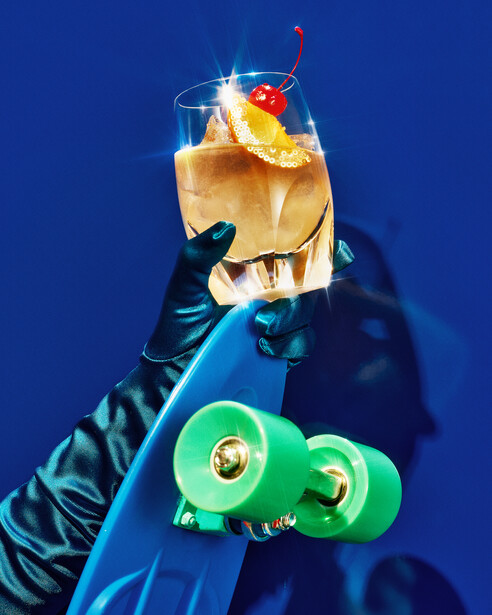 Hand holding a vibrant cocktail with ice, cherry, and citrus garnish against a deep blue background.