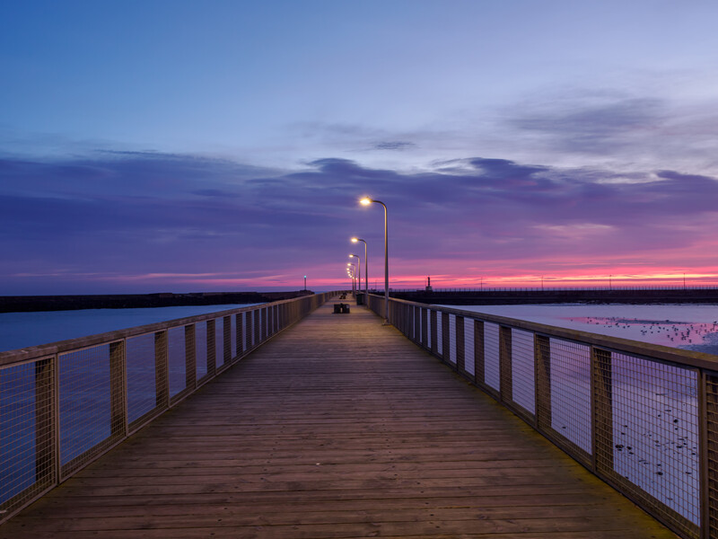 Wooden pier extending into calm water at twilight with street lamps illuminated under a purple and pink sky.