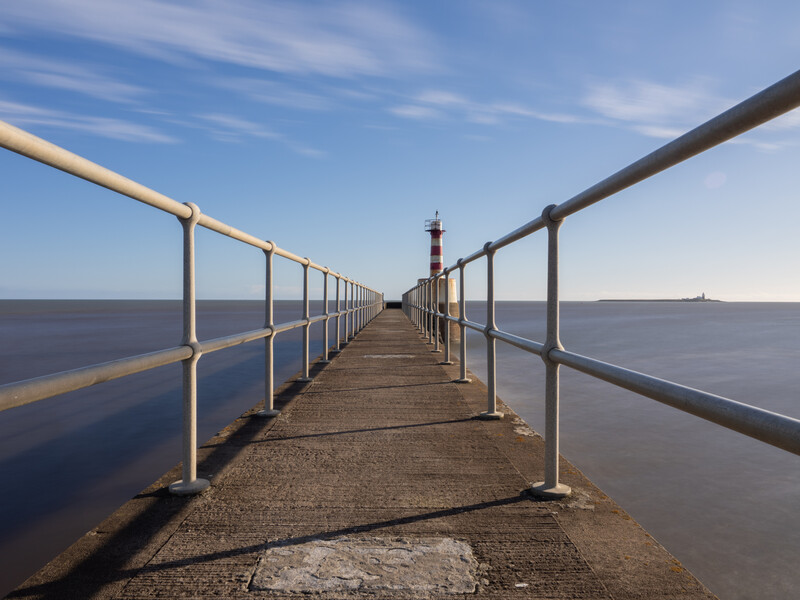 Long wooden pier with metal railings extending toward a red and white lighthouse under blue sky.