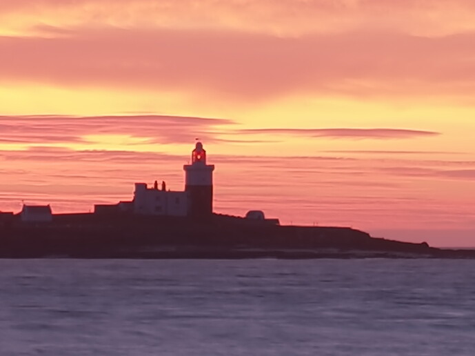 Silhouetted lighthouse on rocky coastline at golden hour sunset with pink and yellow sky.