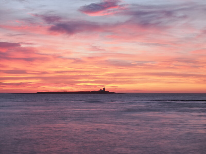 Distant lighthouse silhouetted against a vivid sunset sky with pink, orange, and purple clouds reflected in calm water.