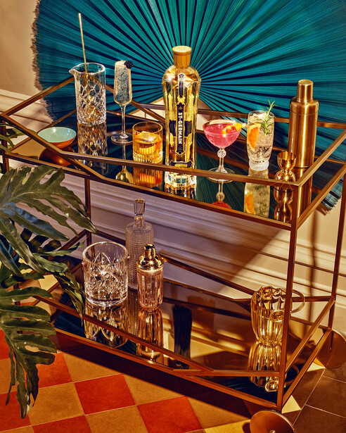 Two-tier bar cart with golden bottles, cocktails in various glassware, and decorative items against a teal fan backdrop.