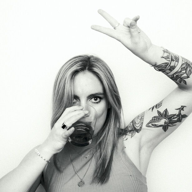 Black and white portrait of a woman with long hair and arm tattoos, drinking from a mug with her eyes closed.