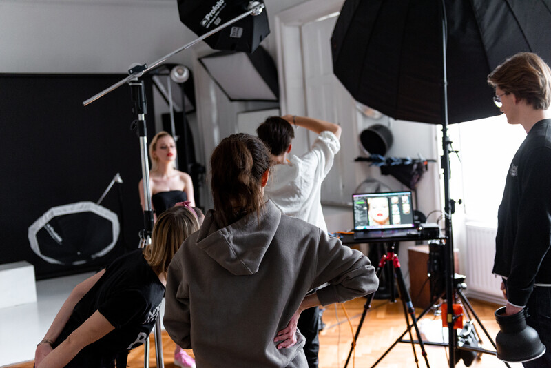 Photography studio setup with overhead softbox lighting, model posing, and crew members adjusting equipment and monitoring on laptop.
