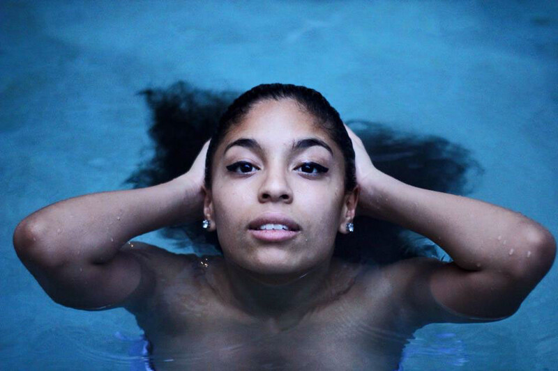 Woman in water with hands behind head, looking directly at camera against blue background.