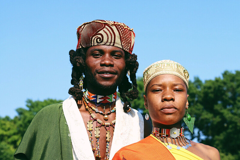 Two people wearing traditional African clothing and jewelry pose against a clear blue sky.