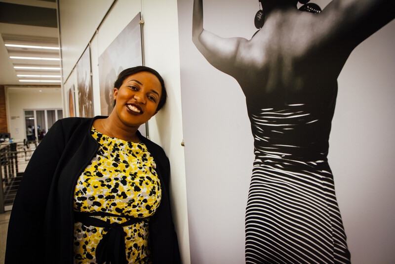 Woman in yellow patterned top smiling next to large-scale portrait photograph on white gallery wall.