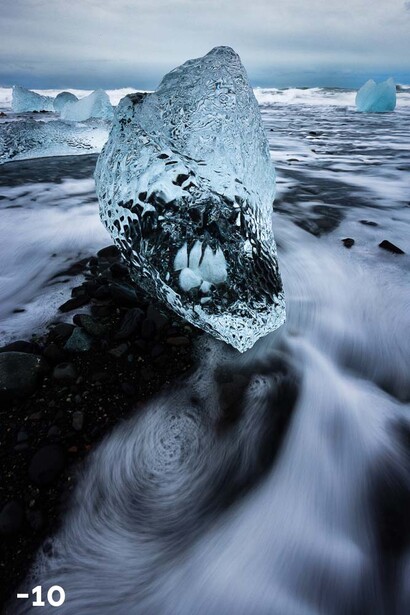 Ice-covered rock jutting from turbulent seawater with snow-capped mountains in the background.