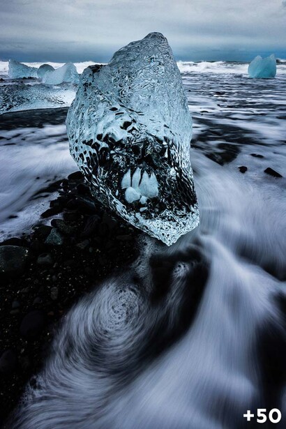 Ice-covered rock with ocean waves flowing around it on a dark volcanic beach.