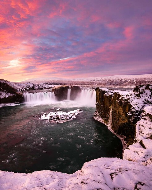 Frozen waterfall and river landscape under vibrant pink and purple sunset sky.