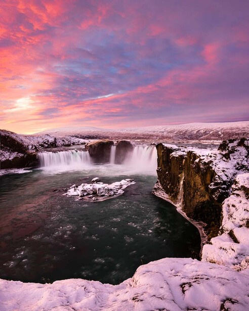 Frozen waterfall at sunset with pink and purple sky reflected in icy water below.
