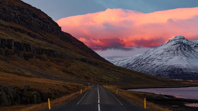 Straight road stretching toward snow-capped mountains under a dramatic pink and orange sunset sky.