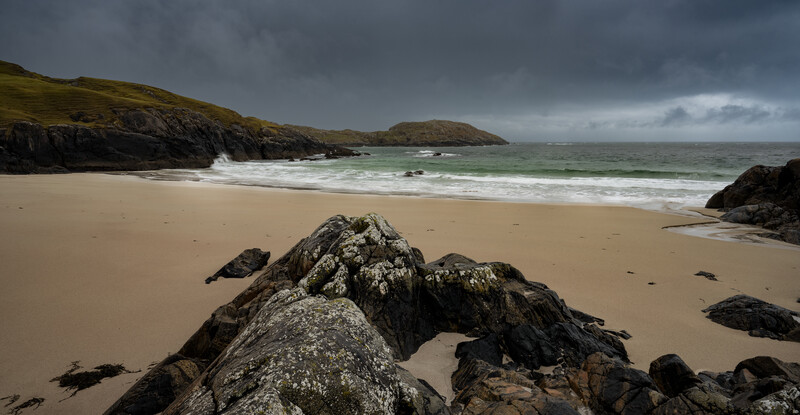 Rocky outcrop on sandy beach with rolling waves and dramatic cloudy sky over distant headlands.