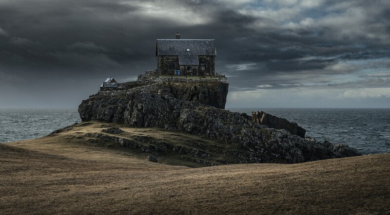 A small stone cottage perched atop a rocky outcrop jutting from a sandy beach under dramatic storm clouds.