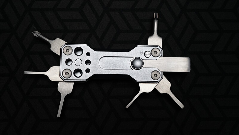 Overhead view of a multi-tool or portable device with symmetrical metallic construction against a dark geometric patterned background.