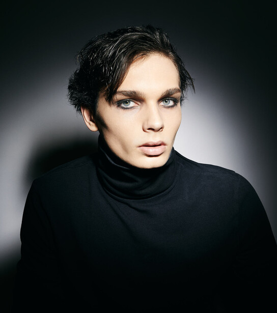 Person with short dark hair wearing a black turtleneck against a dark gradient background.