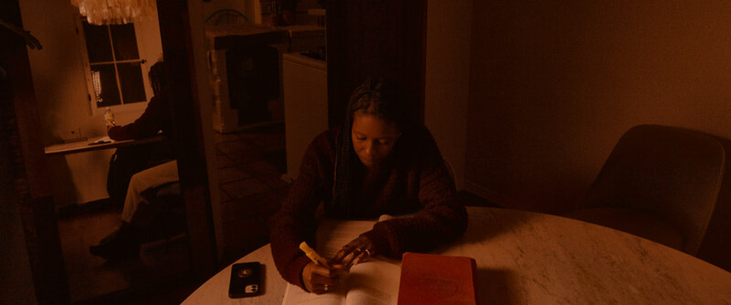 Woman working at a wooden table in warm, dim indoor lighting, focused on a task with papers and objects in front of her.