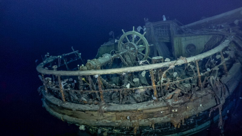 Deteriorated shipwreck on dark ocean floor with visible ship's wheel and wooden hull structures.