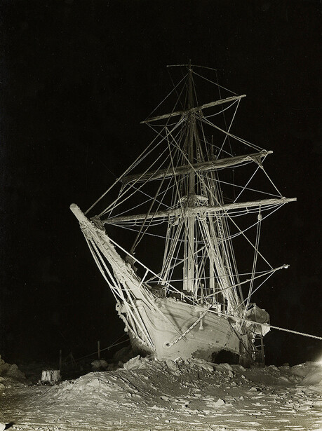 Tall ship with full rigging tilted dramatically in shallow water against dark sky.