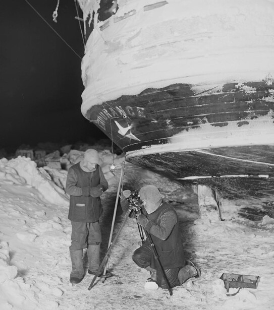Two people in military winter gear standing beside a snow-covered aircraft fuselage.