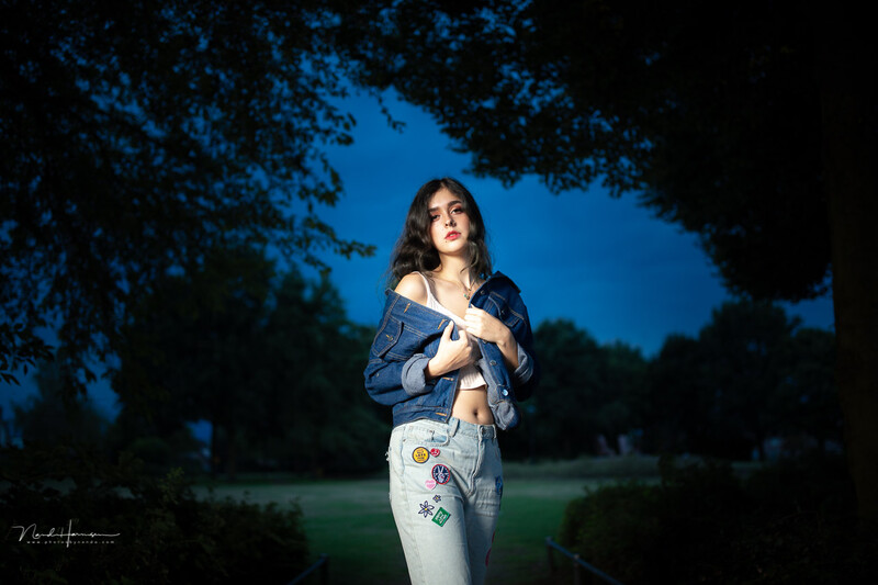 Woman in denim jacket and white pants standing in a dark forest clearing under blue twilight sky.