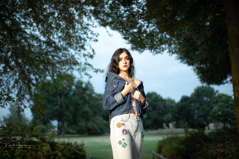 Woman in blazer and embroidered pants standing beneath tree canopy in green field.