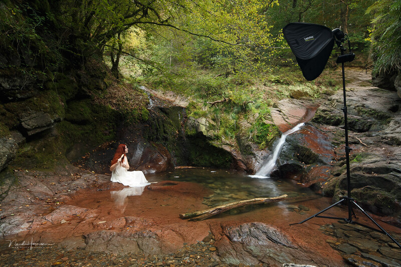 Woman in white dress wading in shallow stream surrounded by forest, with professional lighting setup visible.