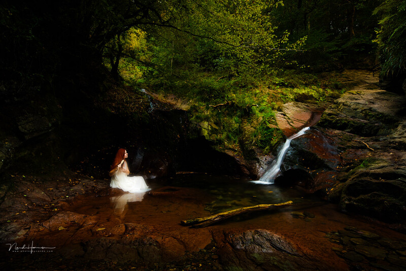 Woman in white dress wading in shallow stream beneath rock overhang with cascading waterfall.