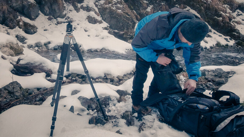 Mountaineer in blue jacket adjusting gear beside tripod on snowy alpine terrain.