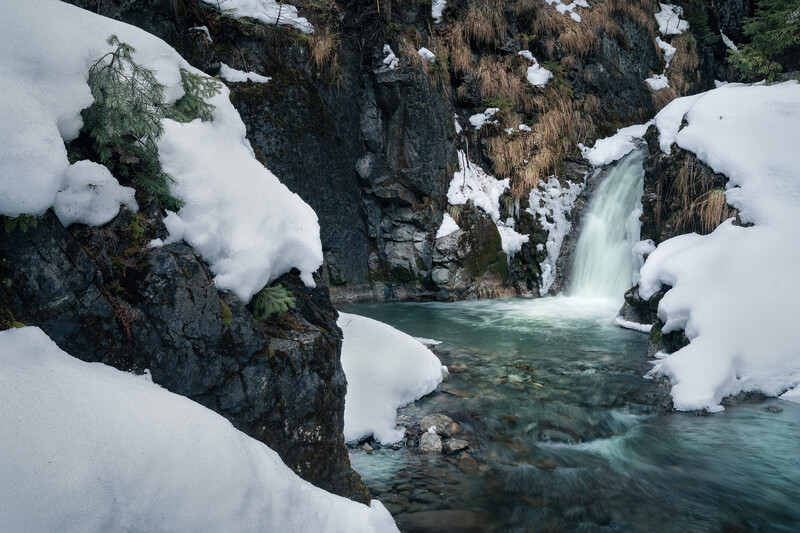 Frozen waterfall cascades between snow-covered rock formations in a winter landscape.