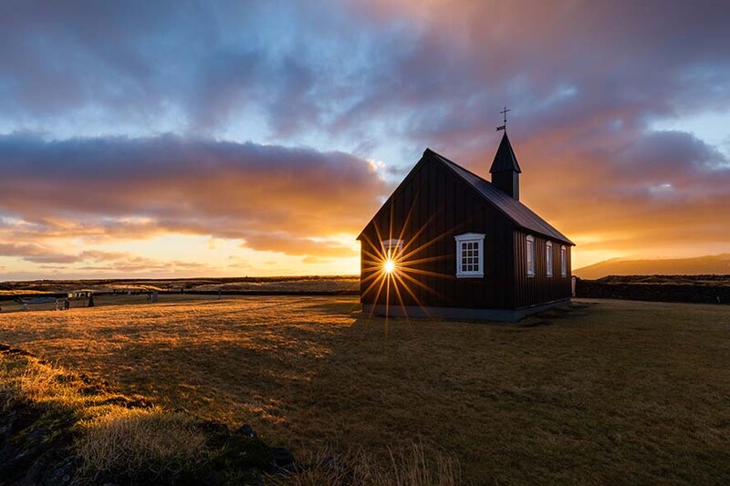 Small dark wooden church with steeple silhouetted against dramatic golden and blue sunset sky on barren landscape.