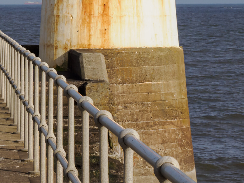 Weathered white lighthouse tower with rust stains beside a wooden railing overlooking open water.