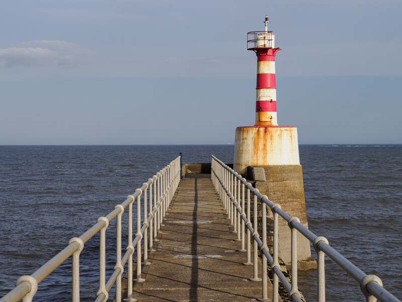 Red and white striped lighthouse at the end of a wooden pier extending into calm sea.