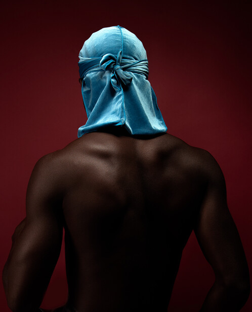Shirtless figure with muscular build wearing a bright blue fabric hood covering their head against a deep red background.