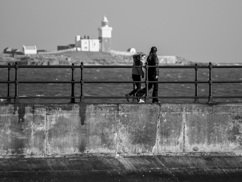 Two figures standing at a railing on a pier, gazing toward a distant lighthouse across the water.