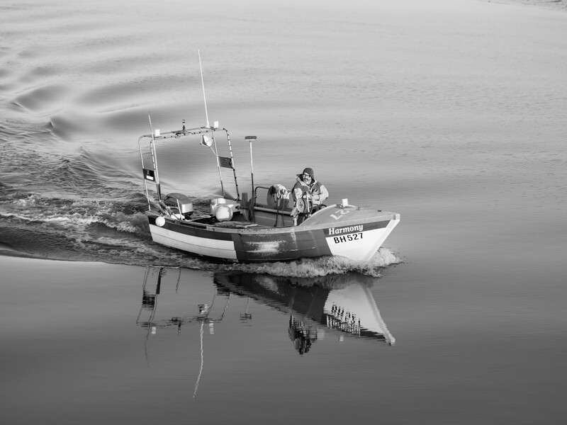 Small fishing boat with registration number visible, photographed in black and white with mirror-like water reflection.