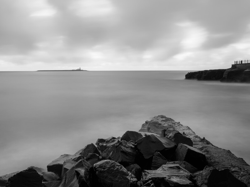 Rocky shoreline in foreground with smooth, misty water and distant headland under overcast sky.
