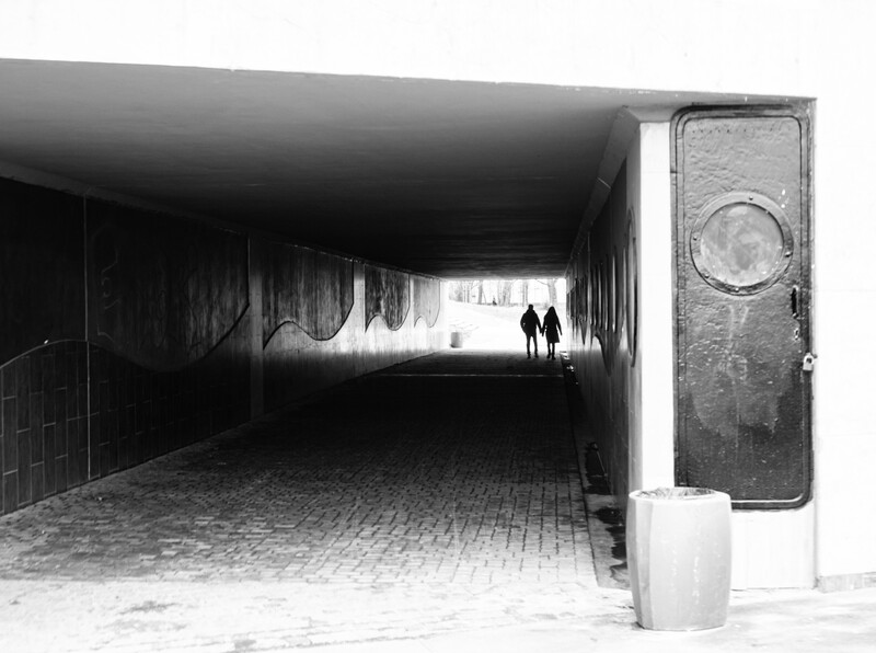 Two silhouetted figures visible through the far end of a dark concrete underpass, rendered in high-contrast black and white.