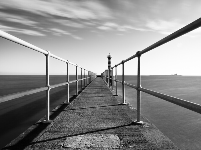 Symmetrical perspective view down a long concrete pier with metal railings extending toward a distant lighthouse.
