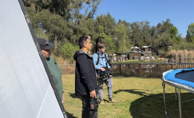 Two people standing in a backyard with camera equipment, observing a photography or video setup near a trampoline and waterfront.