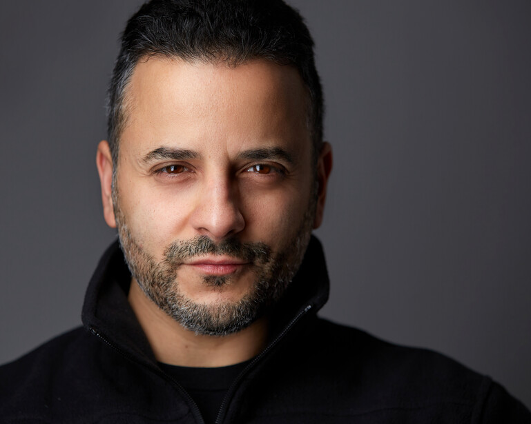 Man with dark hair and beard looking directly at camera against neutral gray background.