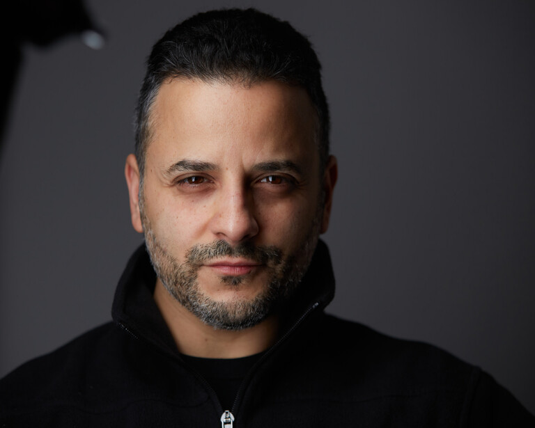 Man with dark hair and beard wearing black hoodie against neutral gray background.