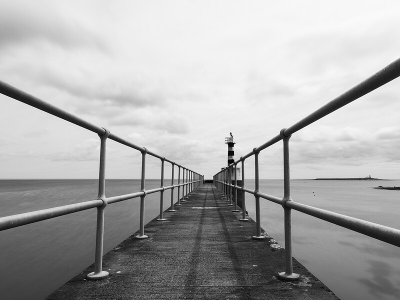 Black and white photograph of a long pier extending toward the horizon with metal railings on both sides.