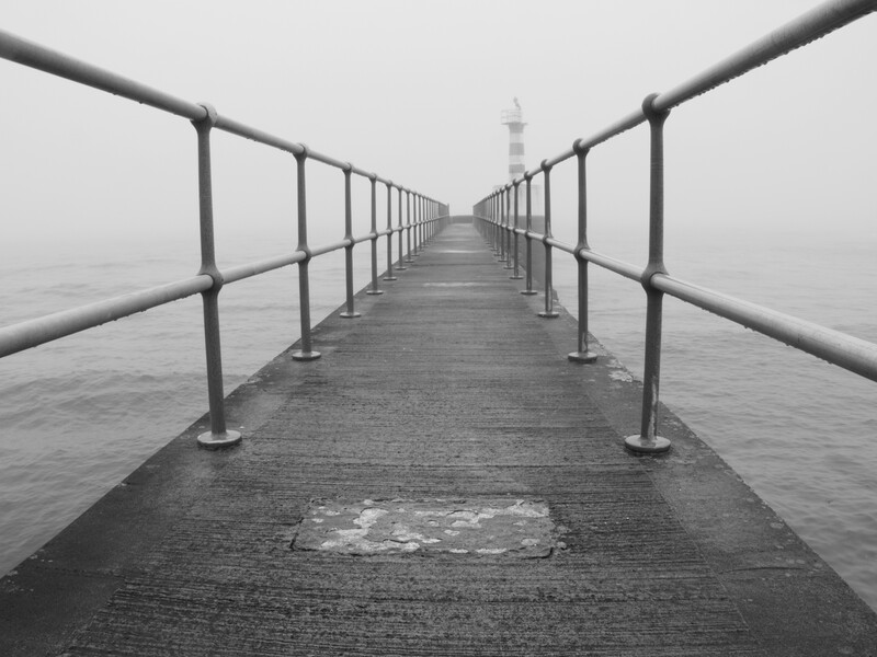 Long wooden pier extending into fog with metal railings on both sides and a distant structure at the end.