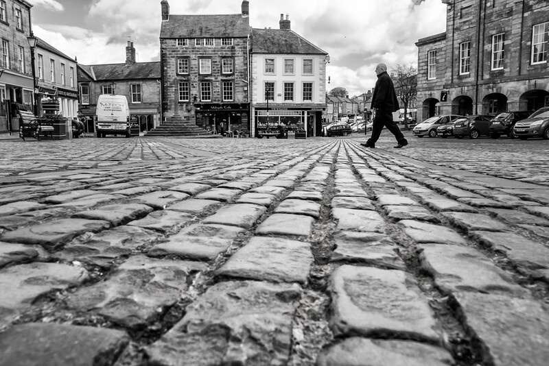 Perspective view of cobblestone plaza with figure walking across radial stone pattern toward historic buildings.