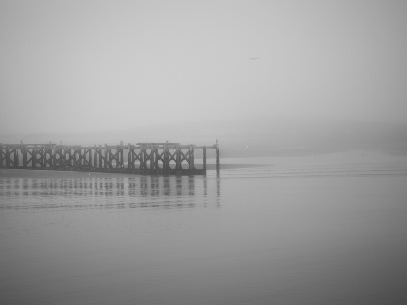 A wooden pier extends into calm, misty water under overcast skies, its reflection mirrored perfectly below.