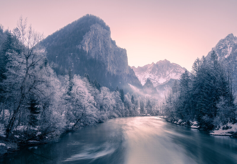 Frost-covered trees frame a misty river valley between steep mountain peaks at dawn.